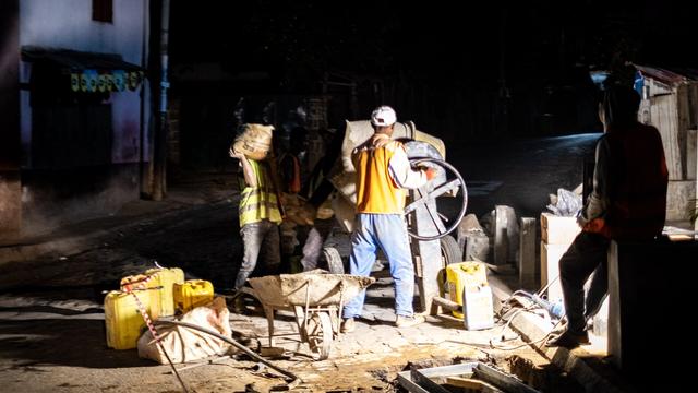 Des ouvriers posant une chambre télécom, de nuit, à un carrefour, à la lumière d'un puissant projecteur.