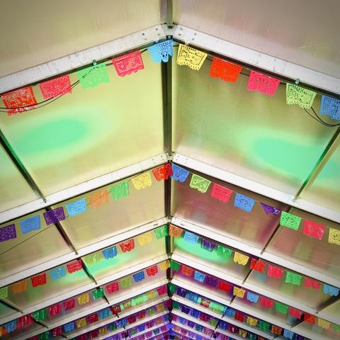 Rows of colorful papel picado banners drape from the vaulted ceiling beams of a large white, metal-framed events tent, as green spotlights shine on every other ceiling section in between.