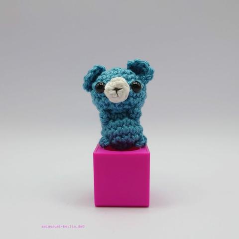 ein pfaufarbener gehäkelter Bär auf einem pinkfarbenen Block - a peacock-coloured crocheted bear on a pink block