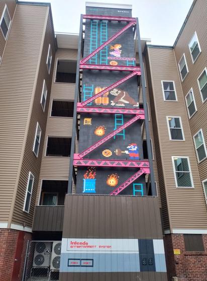 Pixel-art mural styled after the classic Donkey Kong arcade game, painted on the vertical stairwell of a residential building in Concord, New Hampshire. The artwork features Mario, Princess Peach, Donkey Kong, and fireballs across multiple levels with ladders and pink girders, mimicking the game's layout. The bottom of the mural includes a design resembling a Nintendo Entertainment System (NES) console labeled “Intendo Entertainment System” with power and reset buttons. The retro video game theme is seamlessly integrated into the urban architecture, turning the building into a nostalgic tribute to early gaming culture.