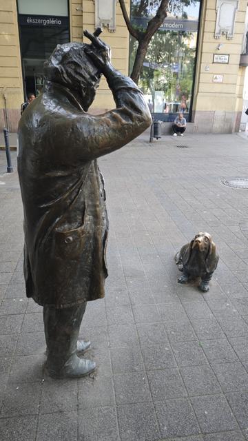 A bronze statue of the star of the TV crime series lieutenant Columbo in a typical pose, scratching his head with the end of a cigar in his fingers. Seen from the side. In front of him an occasional character, a dog, I guess the basset breed, sitting and looking up to him. Seen from the front. The whole scene is on the street.