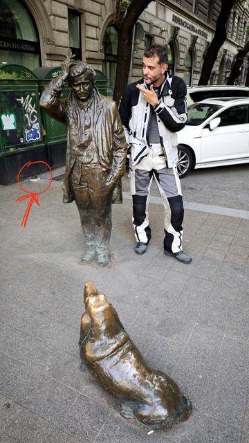 The same statues, this time Columbo seen from the front, the dog from the back. Next to the lieutenant stands a person (in white/black textile motocycle pants and jacket, making a thinking pose to like they are trying to solve the case together. Behind them on the pavement is a small spot, circled in red with an arrow added to the photo.