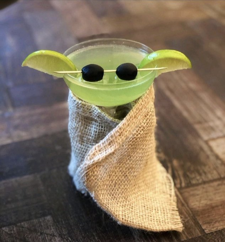 Ein flaches Cocktailglas mit zwei schwarzen Oliven, zwei halben Zitronen und einem Bastumhang. Die Kombiation sieht aus wie Yoda.