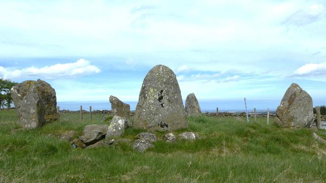 Five of the six chunky standing stones forming a circle in a grass field. The sixth stone is hidden behind the front one.