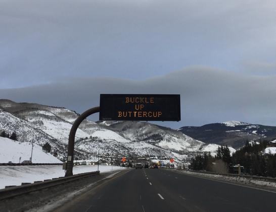 Over two lanes of a four-lane, divided highway along a snowy, high-mountain pass, an informative digital sign reads Buckle Up Buttercup