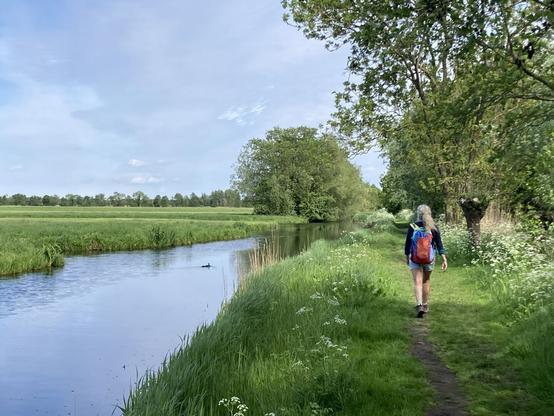 Vrouw op de rug gezien loopt over graspad langs water.