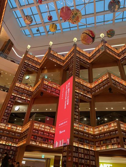 One side of the main library space. The walls go up four stories, with gaps like windows where people on other floors can look down. The walls go in and out in a triangle pattern and are entirely bookshelves loaded with books. Everything is lit in warm colours.
