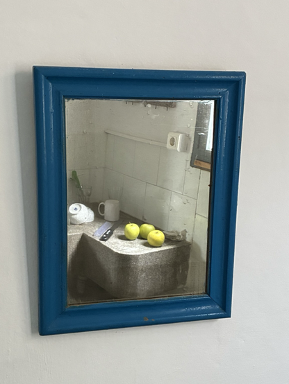 Auf einer Terrazzo-Spüle: Drei hervorragend beleuchtete grüne Äpfel, zum Trocknen hin gestellte Schüsseln und ein Becher und eine Microplane-Reibe, all dies in einem Spiegel in blauem Holzrahmen an einer weiß gestrichenen Wand.