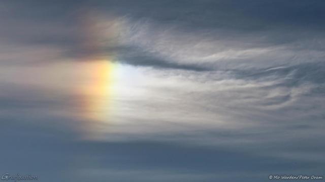 A colour photo of wispy white clouds on a darker greyish background. Left of centre is a stripe of beautiful rainbow colour visible from red-orange to pale yellow with hints of the other colours on each side.