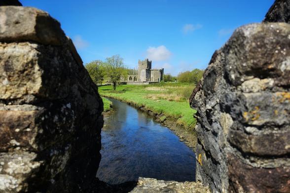 A stone crenulation frames a view of Tintern Abbey standing in a green field beside a flowing river under a bright blue sky with a few clouds.