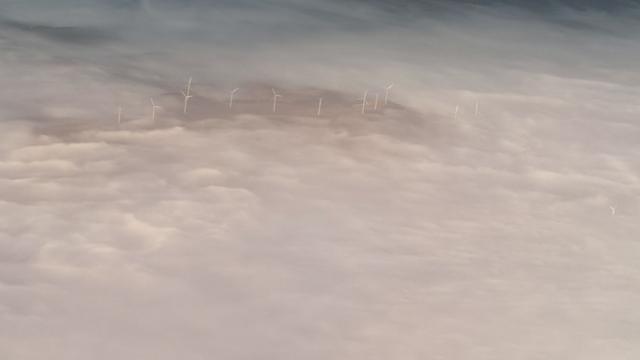 Photograph looking down out the window of a plane. A few wind turbines appear as tiny towers peeking through low cloud.  It is early morning and the sun is colouring the clouds a shining silver with patches of pink and blue.