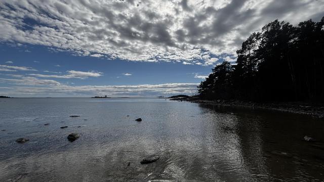 View over the water with some cloud coverage, trees in the right, small isalnds, and some stones sticking up out of the shallow water.