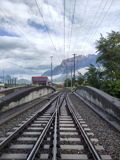 Foto. Betonbrücke mit einer Weiche, im Hintergrund Berge und ein roter Zug.