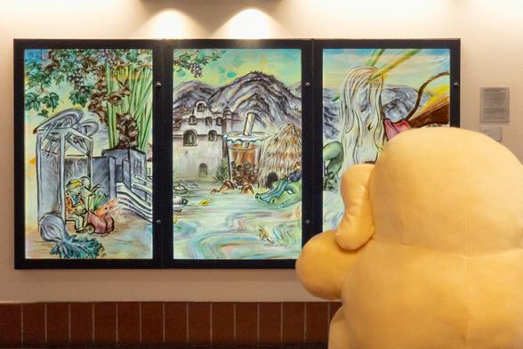 Photo of the Creature Mastodon stuffed toy in front of posters celebrating the city of San Gabriel on a warmly lit wall. The Creature is in the foreground, its back to the camera as it stares forward at Sandra Low's “San Gabriel” artwork for LA Metro's “Through the Eyes of Artists” poster series in the Passageway Art Gallery of Union Station Los Angeles.