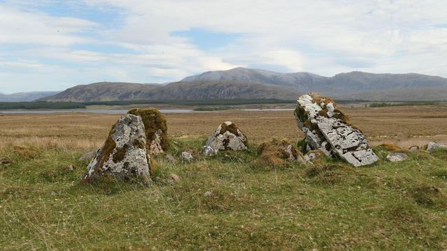 The cairn material is mostly gone from the chambered cairn but the orthostats of the chamber still remain. Open landscape to the big hills beyond.