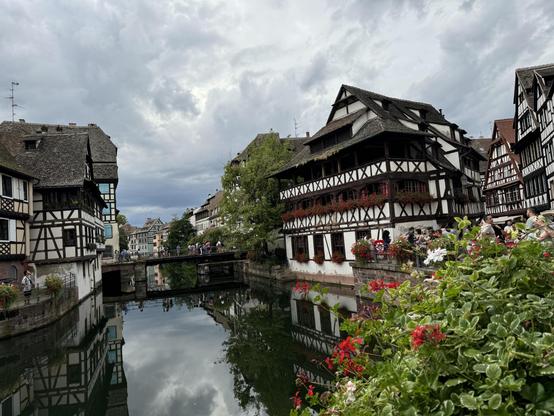 The image depicts a picturesque scene of what looks like a historic European town with traditional half-timbered houses lining a canal. The buildings reflect in the water, and there are flowers and greenery along the canal's edge. A cloudy sky adds to the scenic atmosphere, and there are people visible on a bridge in the background.