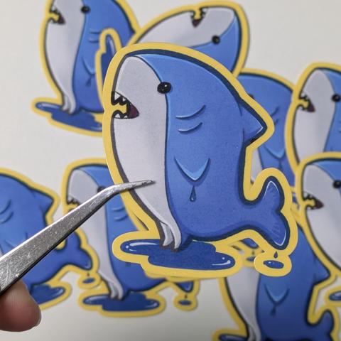 Stickers of the ffxiv minion major general, a little shark with legs. He's standing in a puddle of water, and water is dripping off its tail.

A different perspective, the sticker is held up by pincers