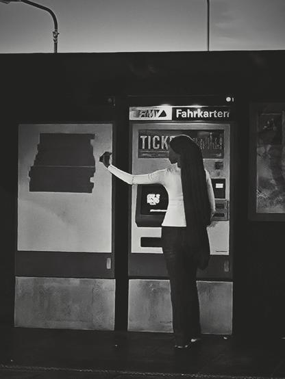 The black and one image shows a girl taking a selfie while standing in front of a ticket machine. She's wearing a light-colored, long-sleeved shirt and dark pants. A scarf is tied around her head and falls down her back all the way to her thighs. Her right arm is extended, holding a phone pointed towards herself. The ticket machine has the words "FAHRKARTEN" and "TICKET" visible on it. To the left of the machine is a blank panel, and to the right, part of another panel is visible. A light fixture is present in the upper left of the frame.