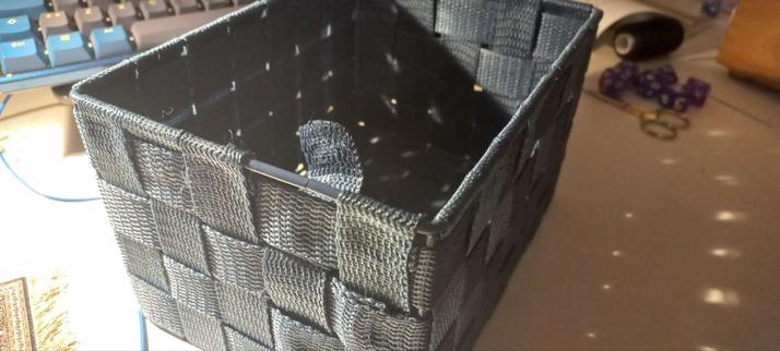 Una cesta hecha de cintas de tejido sintético. No están cosidas sino termoselladas, y una de ellas se ha despegado del marco metálico. Al fondo pueden verse unos dados de rol, un teclado mecánico y enseres de costura. La luz es artificial, y mala para una foto; es una imagen nocturna.