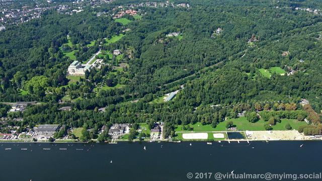 An aerial photo of the coastline of the Baldeneysee in the direction of Villa Hügel, which can be seen in the middle left next to many trees. There are some small sailing boats in the lake.