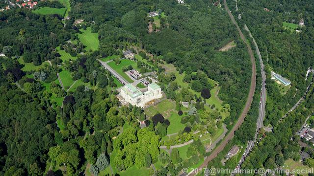 An aerial photo of the Villa Hügel and the Hügelpark, sourrounded by many trees. A trainline with a stop in front of the main building can be seen in the bottom right.