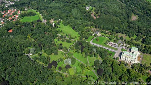 An aerial photo of the Villa Hügel and the Hügelpark, sourrounded by many trees. You can see the parking lot and the building of the Krupp foundation in front of the main building.