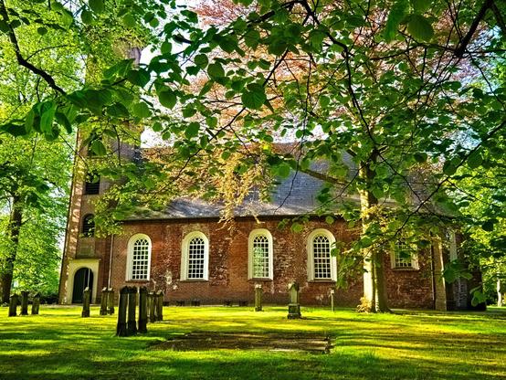 A pretty church with a lawn in front of it. There are various gravestones on it. Green leaves/trees can be seen everywhere. This reformed church was built in the 13th century and served as a burial place for members of the von Wedel family. At the end of the 13th century, the church was built with bricks in a monastery format as a hall church. In the 15th century, the cross-vaulted choir was added, extending the nave by 5.5 metres to the east across its entire width. A tower can be seen on the left, followed by the nave with white-framed arched windows.