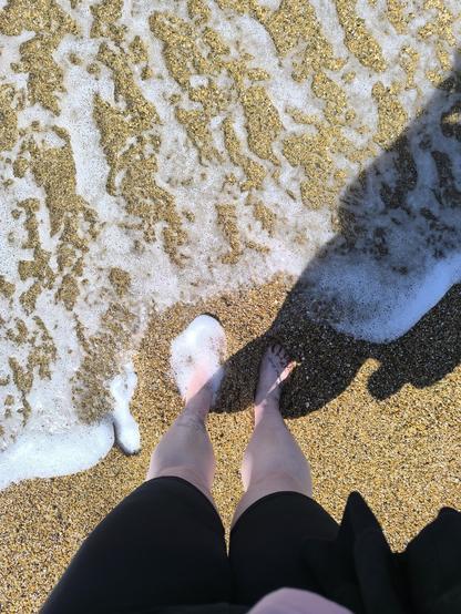 Ich stehe am Strand und fotografiere meine Füße. Das Foto zeigt also den Sand, in der unteren Hälfte sind meine Beine. Ich trage eine schwarze Leggings bis zu Knien, meine nackten Füße sind im Sand eingegraben. Gerade kommt eine Mini Welle, der Schaum ist über die obere Hälfte des Bildes verteilt.