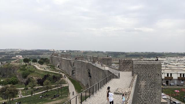 A huge fortress wall with a park on the inside and houses on the outside. Some people are promenading on the wall.