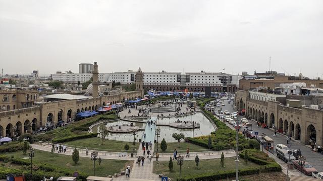 The city park of Hewlêr photographed from the citadel. People are relaxing next to fountains, the streets are vivid and on the right side, there's the bazar.