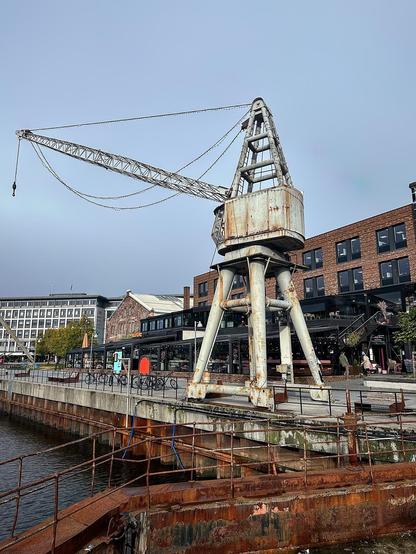 This image features a large, old crane situated by a waterfront. The crane, showing signs of rust and wear, stands tall with its boom extended out over the water. In the background, you can see modern buildings, possibly containing restaurants or shops, with outdoor seating areas. The setting suggests a renovated industrial area that has been repurposed for commercial or recreational use. There is also a dock visible in the foreground, and the overall atmosphere is of an urban waterfront that blends historical industrial elements with contemporary urban life.