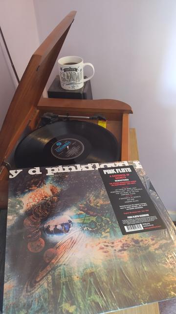 Record player with a 12" black vinyl record.
In the foreground is an album cover with a small photograph of the band inset into an abstract design. A promotional sticker is fixed to the right hand side.
Behind the player is a mug of coffee on a speaker.