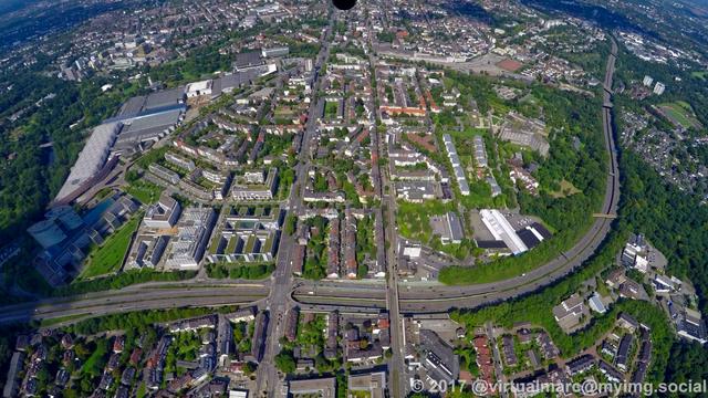 An aerial photo of Essen Rüttenscheid. You can see multiple living quaters sourrounded by many trees. There is a freeway going through the image (the A52). At the top left you can see the Essen fair and the Grugapark.
