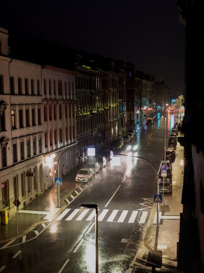 Blick von oben auf eine nasse Strasse bei Nacht. Die Lichter reflektieren sich auf der Straße und den Häuserwänden.