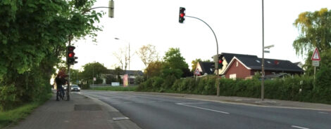Ein Junge steht an einer Anforderungsampel, um die Straße zu queren. Die Ampel ist gerade auf Orange umgesprungen. Zwei PKW sind noch ca. 15 bis 20 Meter von der Haltelinie der Ampel entfernt.
