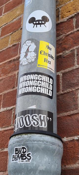 Portrait oriented close up of part of a metal drain pipe with several stickers against a red brick wall. From top to bottom:

An oval white sticker with a black flying saucer with four windows, four legs and Mickey Mouse ears

A white, crackled, mostly gone, illegible sticker on the left of a portrait oriented, left side yellow, right side white sticker saying Ave Christus Rex in a germanic looking font with an instagramlogo followed by jongerenvoorlambertus (youthforlambertus) in small font below it

A landscape oriented sticker with wrongchild in white letters three times from top to bottom on a black background.

A landscape oriented sticker with the word woosh in white outlined black letters tilted to the left in white quotation marks on a black background.

A small sticker cut so the ouline follows the words. It has the word Bud above the word Bombs in a graffiti marker style with the instagram logo followed by bud__bombs below it