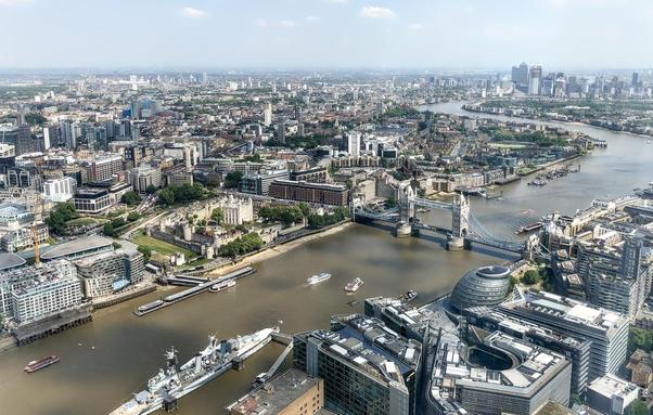 This image shows a sweeping aerial view of London, featuring several notable landmarks. On the right, you can see the Tower Bridge, a famous bascule and suspension bridge over the River Thames. To the left of the Tower Bridge is the historic Tower of London, a historic castle that has played various roles, including a royal palace, a prison, and a treasury. Below and slightly to the right of the Tower of London is a distinctive modern building known as City Hall, which has a unique, rounded design. The photograph captures the Thames snaking through the city, with numerous buildings of various styles and eras lining its banks, illustrating the architectural diversity and historical layers of London.