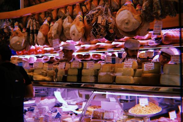 A counter full of cheeses and ham with two works behind it, only their heads are visible.