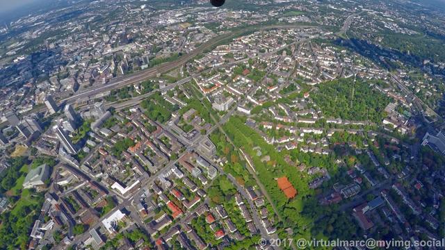 An aerial photo of the centrum of essen. You can see the east cementry on the right. Essen central station with the skyline of Essen on the left. Multiple living quaters and many trees are all around.