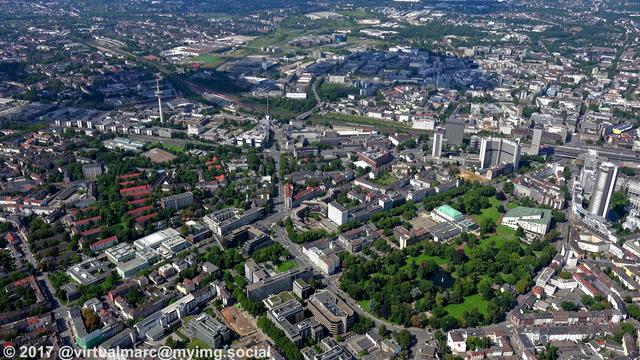 An aerial photo of the center of essen. You can see Essen central station and the innogy Headquaters (former RWE tower) on the right. The Aalto-Theatre and the Philharmonic Essen can be seen together with their park in the bottom right. The telecomincations tower can be seen in the rop left. Around are some business and living quaters with trees next to them.