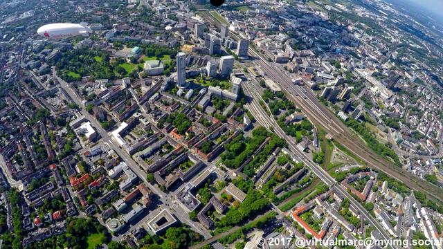 An aerial photo of the center of essen. You can see the main freeway (the A40, ruhr express way, Ruhrschnellweg), the Essen central station, multiple living and working quaters sourrounded by trees, the innogy Headquaters (former RWE tower), the Aalto-Theatre and the Philharmonic Essen. The airship of the WDL Luftschiffgesellschaft mbH is hovering over the city.