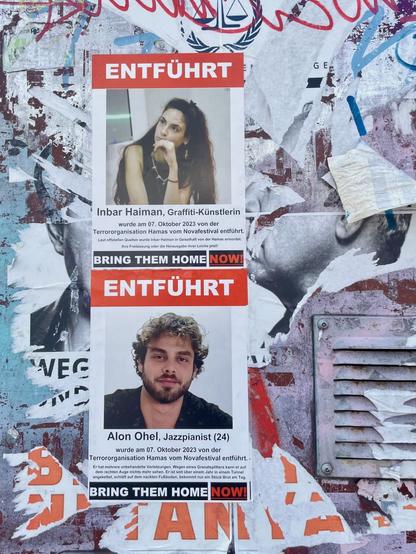 Foto. Ein Teil eines Stromkastens. Auf diesem Tags und abgerissene Plakate. Darüber plakatiert zwei A4-Plakate, jeweils mit der Meldung „ENTFÜHRT“. Jedes Plakat zeigt eine der von der Hamas aus Israel entführten Geiseln. Plakat 1: „Inbar Haiman, Graffiti-Künstlerin wurde am 07. Oktober 2023 von der Terrororganisation Hamas vom Novafestival entführt. Laut offiziellen Quellen wurde Inbar Haiman in Geiselhaft von der Hamas ermordet. Ihre Freilassung oder die Herausgabe ihrer Leiche jetzt! BRING THEM HOME NOW“. Plakat 2:“Alon Ohel, Jazzpianist (24) wurde am 07. Oktober 2023 von der Terrororganisation Hamas vom Novafestival entführt. • Er hat mehrere unbehandelte Verletzungen. Wegen eines Granatsplitters kann er auf dem rechten Auge nichts mehr sehen. Er ist seit über einem Jahr in einem Tunnel angekettet, schläft auf dem nackten Fußboden, bekommt nur ein Stück Brot am Tag. BRING THEM HOME NOW“. Auf beiden Plakaten je ein Porträt. Inbar hat langes dunkles Haar, trägt ein schwarzes ärmelloses Shirt und schaut aus dem Bild heraus. Alon hat dunkles kurzes lockiges Haar und einen Bart, trägt ein schwarzes Oberteil und blickt direkt in die Kamera.