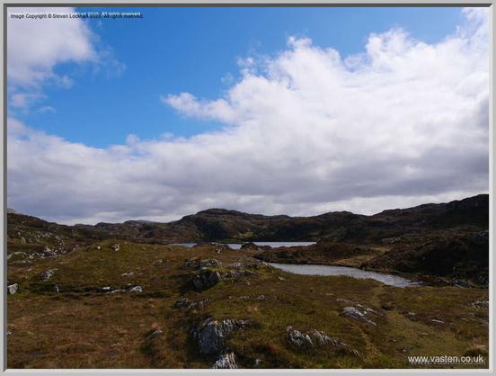 Yellowy vegetation and grey rock on the foreground, a loch in the middle distance and some hills beyond that. Skies are blue and bright white cloud