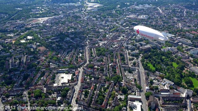 An aerial photo of the center of essen with many trees between the houses. The airship of the WDL Luftschiffgesellschaft mbH is hovering over the city.