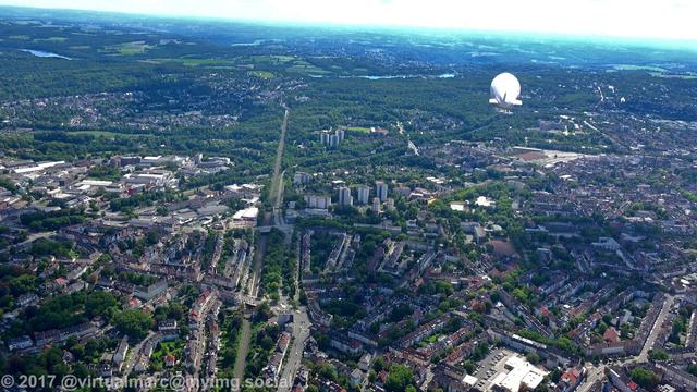 An aerial photo of the center of essen towards the south. You can see many buildings with trees around, The further you look in the distance the less buildings and more trees are showing up. The airship of the WDL Luftschiffgesellschaft mbH is hovering over the city.
