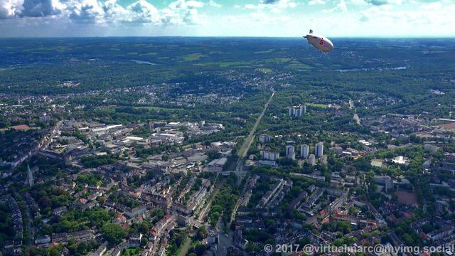 An aerial photo of the center of essen towards the south. You can see many buildings with trees around, The further you look in the distance the less buildings and more trees are showing up. The airship of the WDL Luftschiffgesellschaft mbH is hovering over the city.