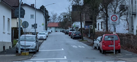 Eine Straße in einer Stadt mit Wohnhäusern rechts und links sowie beidseitigen Gehwegen. Die Straße ist auf beiden Seiten zugeparkt, wobei einige Fahrzeuge mit einer Spur bereits auf dem Gehweg stehen, da die Parkbuchten recht schmal sind.