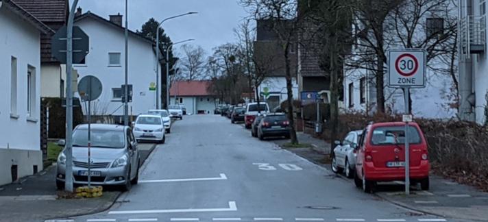 Eine Straße in einer Stadt mit Wohnhäusern rechts und links sowie beidseitigen Gehwegen. Die Straße ist auf beiden Seiten zugeparkt, wobei einige Fahrzeuge mit einer Spur bereits auf dem Gehweg stehen, da die Parkbuchten recht schmal sind.