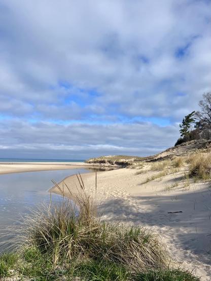 A serene beach scene featuring a sandy shoreline with gentle dunes, patches of grass, and a calm, winding river. The sky is partly cloudy, with blue patches visible. Lake Michigan is seen in the background, creating a tranquil atmosphere.
