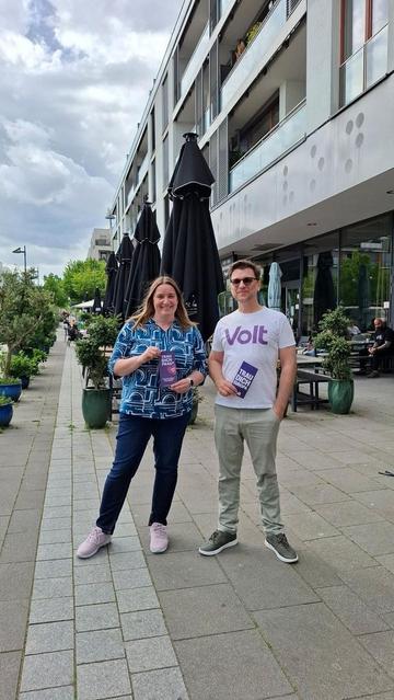 Eine Frau in bunter Bluse und ein Mann mit "Volt"-T-Shirt stehen nebeneinander vor einem modernen Gebäude und lächeln in die Kamera. Beide halten Flyer in der Hand. Auf dem der Frau steht "Trau dich, Frau", auf dem des Mannes "Trau dich Europa". Im Hintergrund sind außerdem (von links nach rechts) zwei Reihen von Pflanzenkübeln, eine Reihe Sonnenschirme und die Tische eines Cafes zu sehen. An einem der Tische sitzt ein Mann, der zu den beiden hinübersieht.
Alle Fluchtlinien weisen zum Horizont, alle vertikalen Linien auf die beiden Zentralfiguren.